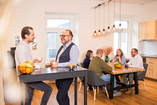 Zwei Männer stehen und unterhalten sich an einem hohen Tisch mit Obst, während vier Personen an einem hölzernen Esstisch in einem modernen, hellen Raum zusammensitzen und sich unterhalten.