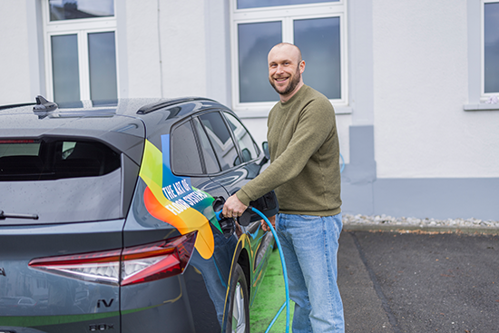 Ein lächelnder Mann steht neben einem grauen Elektroauto und schliesst es an eine Ladestation vor einem weissen Gebäude mit grossen Fenstern an. Das Auto hat einen bunten Aufkleber von UZIN UTZ an der Seite.