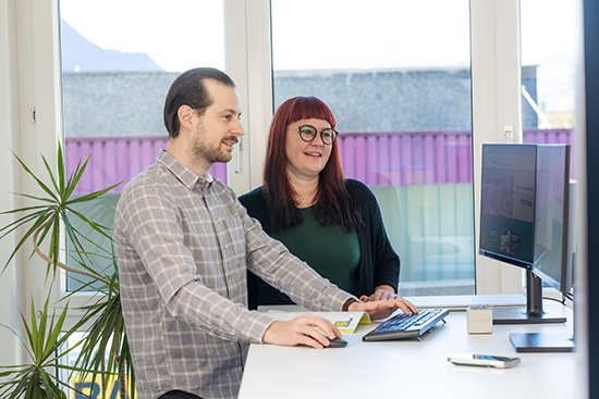 Zwei Mitarbeitende der UZIN UTZ Schweiz stehen an einem Schreibtisch in einem Büro und arbeiten gemeinsam an einem Computer. Der Mann bedient Maus und Tastatur, während die Frau lächelnd neben ihm steht. Im Hintergrund sind eine Pflanze und grosse Fenster zu sehen.